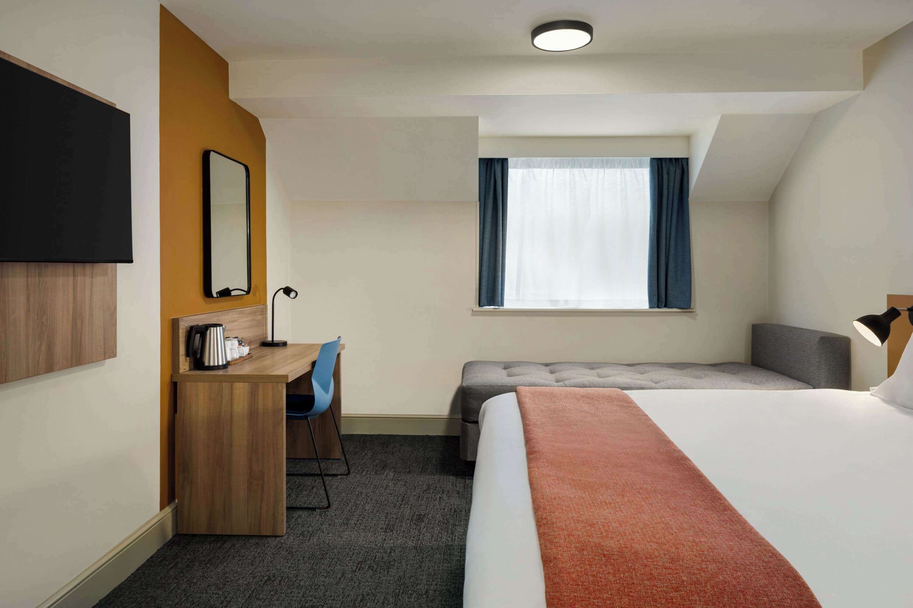 A hotel room with a bed, a desk, a chair, a TV, a window, and a ceiling light.