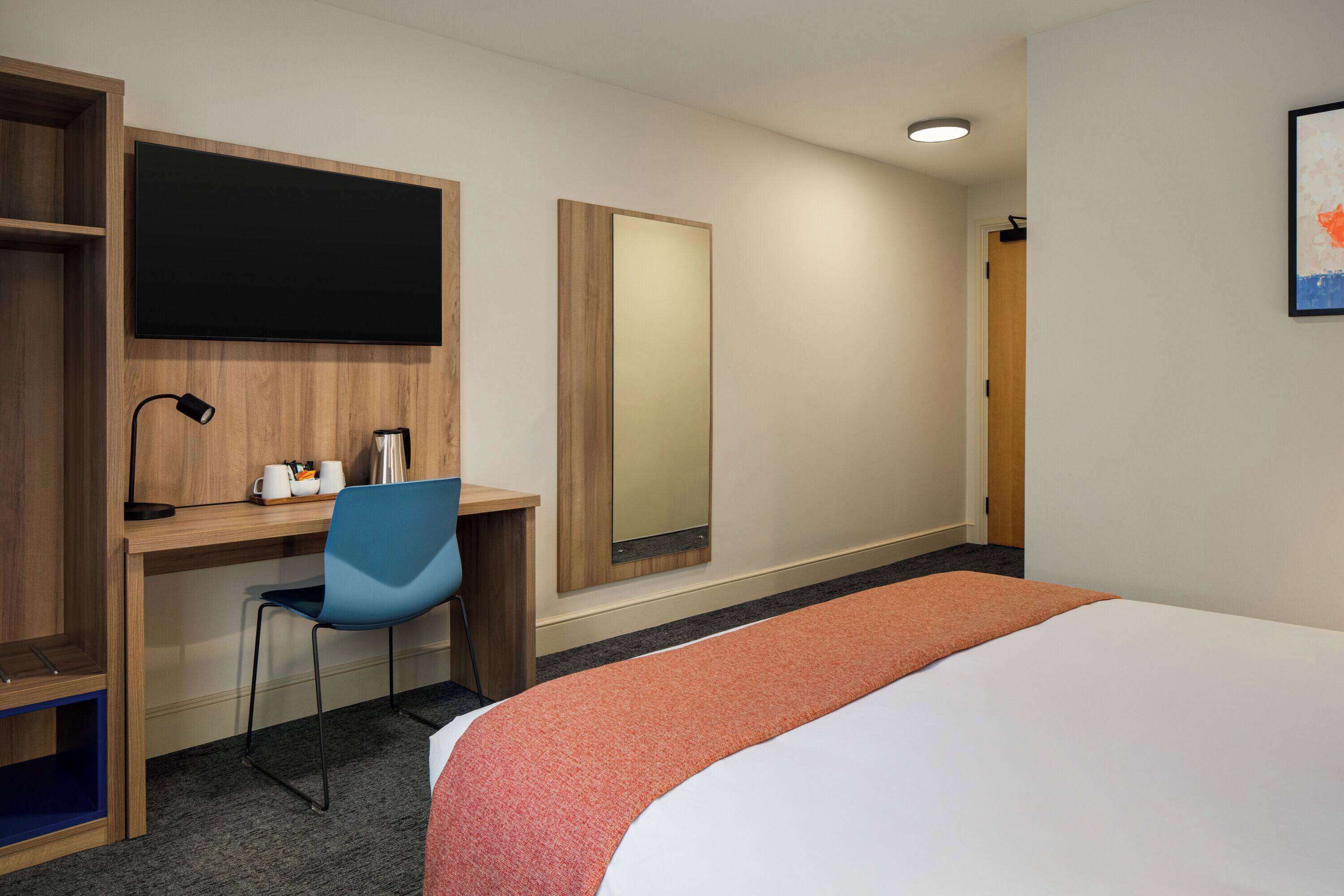 A hotel room with a bed, a desk with a chair, a large mirror, and a bookshelf.