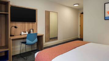 A hotel room with a bed, a desk with a chair, a large mirror, and a bookshelf.