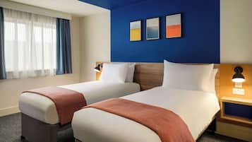 Habitación de hotel con dos camas, un ventanal con cortinas, una pared de acento azul y cuadros enmarcados.