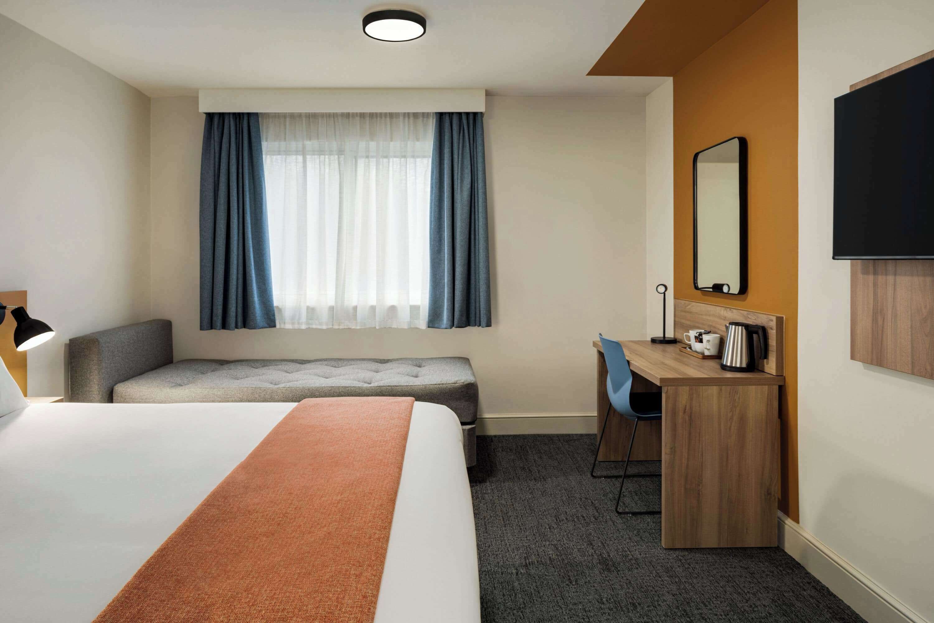 Habitación de hotel con una cama grande, un escritorio con silla, un pequeño sofá, una ventana con cortinas, un televisor de pantalla plana y una cafetera.
