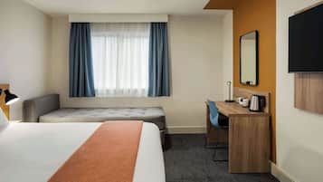 Habitación de hotel con una cama grande, un escritorio con silla, un pequeño sofá, una ventana con cortinas, un televisor de pantalla plana y una cafetera.