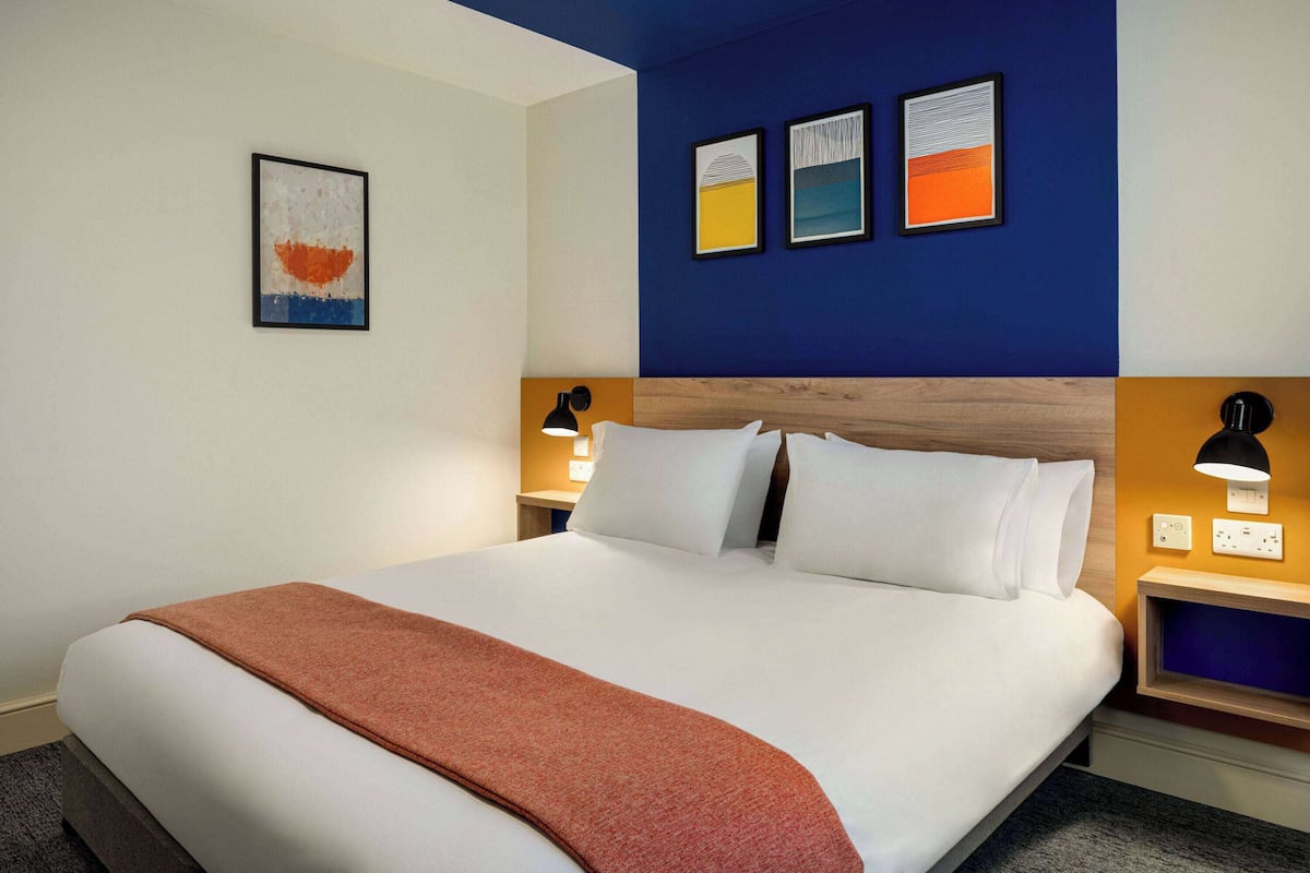 A hotel room with a large bed, a brown throw blanket, two bedside lamps, and artwork on the wall.