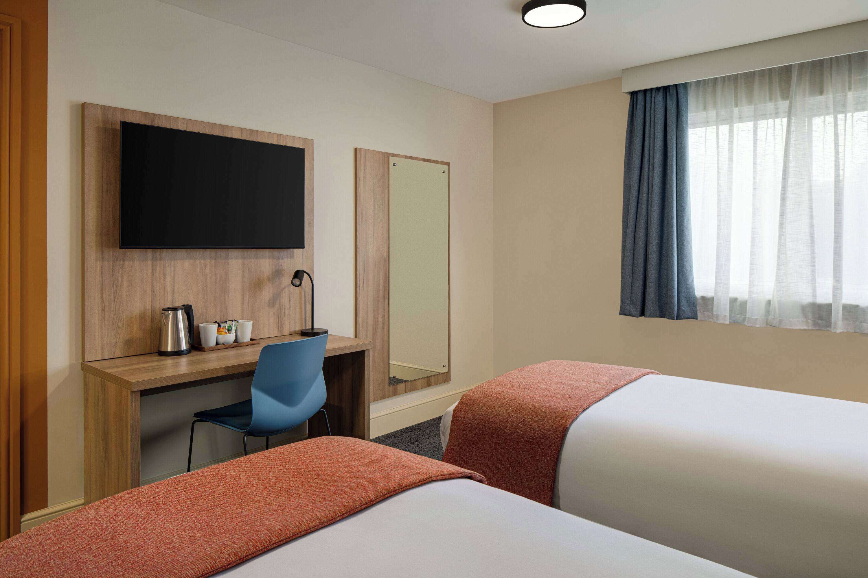 Habitación de hotel con dos camas, un escritorio con silla, televisor de pantalla plana, espejo y ventana con cortinas.