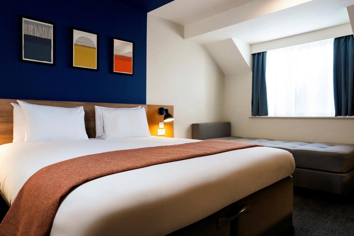 A hotel room with a large bed, a bedside table, a lamp, a window with curtains, and artwork on the wall.
