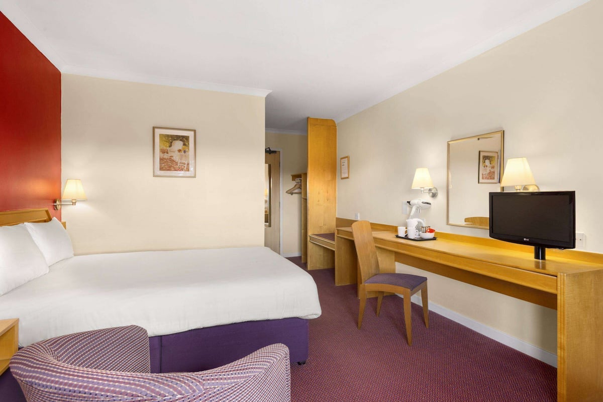 A hotel room with a bed, a desk with a chair, a television, and a small table with a lamp.