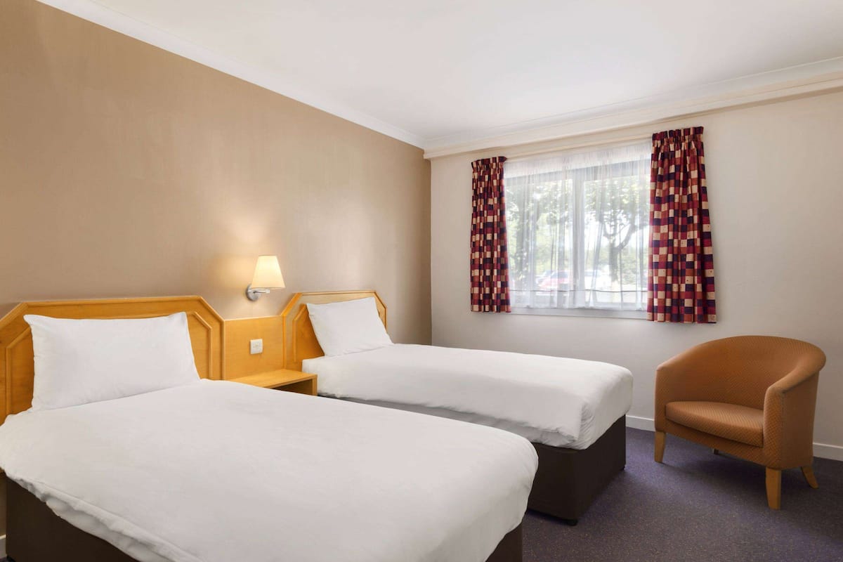 A hotel room with two beds, a chair, a window with curtains, and a lamp.