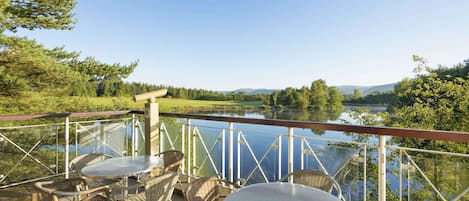 A terrace with wicker chairs and tables overlooking a lake, with a metal railing and a view of the surrounding landscape.
