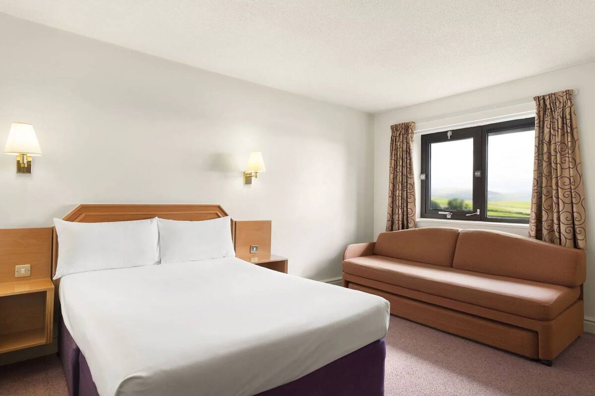A hotel room with a bed, a nightstand, a sofa, two wall-mounted lamps, and a window with curtains.