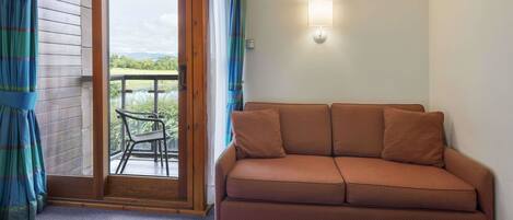 Una habitación con un sofá marrón, un ventanal corredero que da a un espacio exterior con vistas, y una lámpara fijada a la pared.