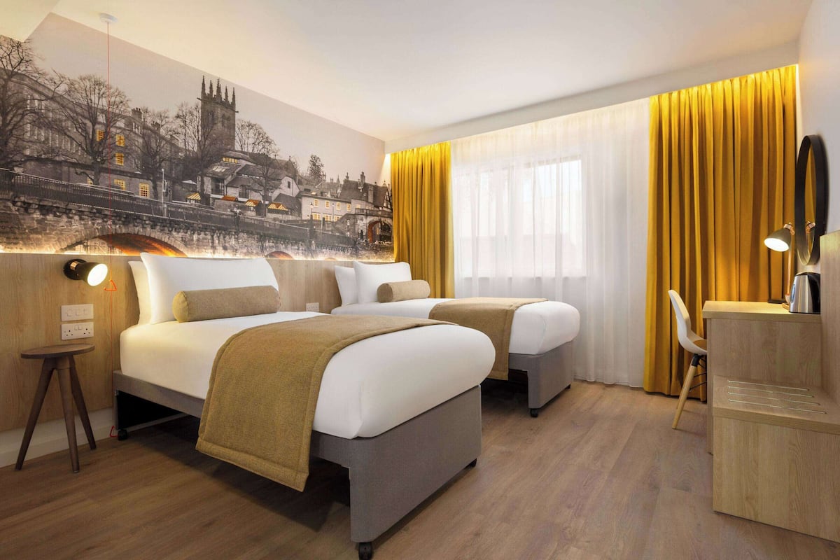 A hotel room with two beds, a desk, a chair, and a mural of a town scene.