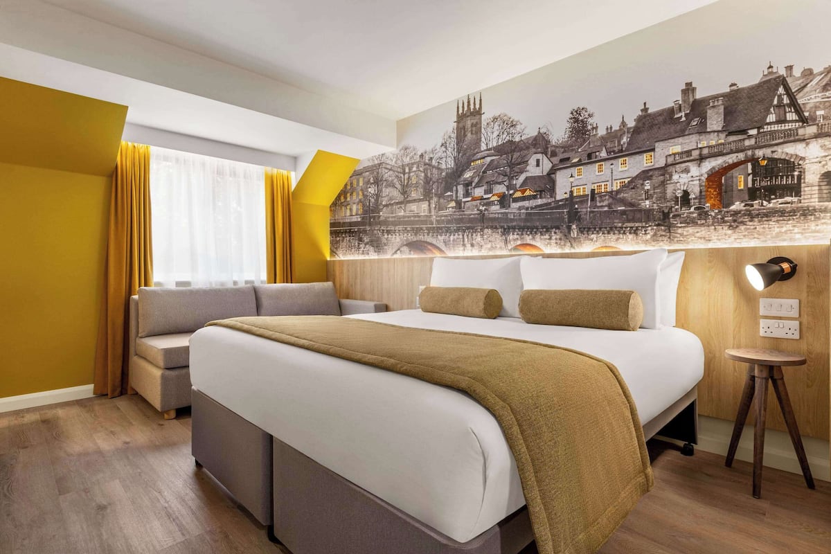 A hotel room with a large bed, a sofa, a small table, and a mural of a historic town on the wall.