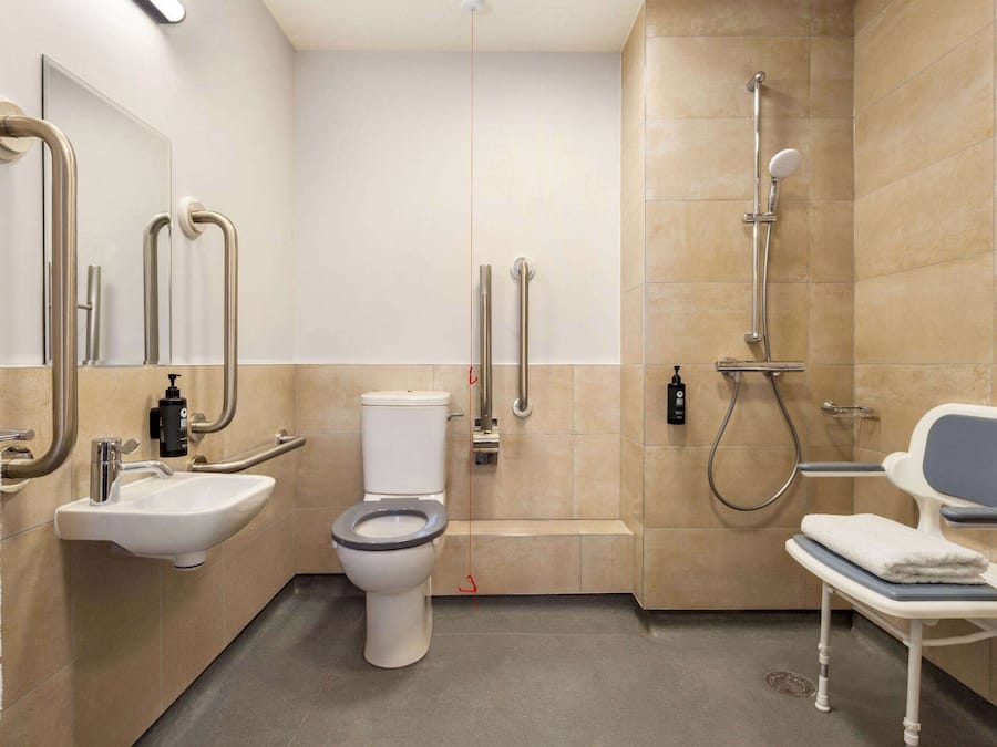 A modern bathroom with a toilet, sink, and shower. It includes grab bars and a wheelchair-accessible bench.