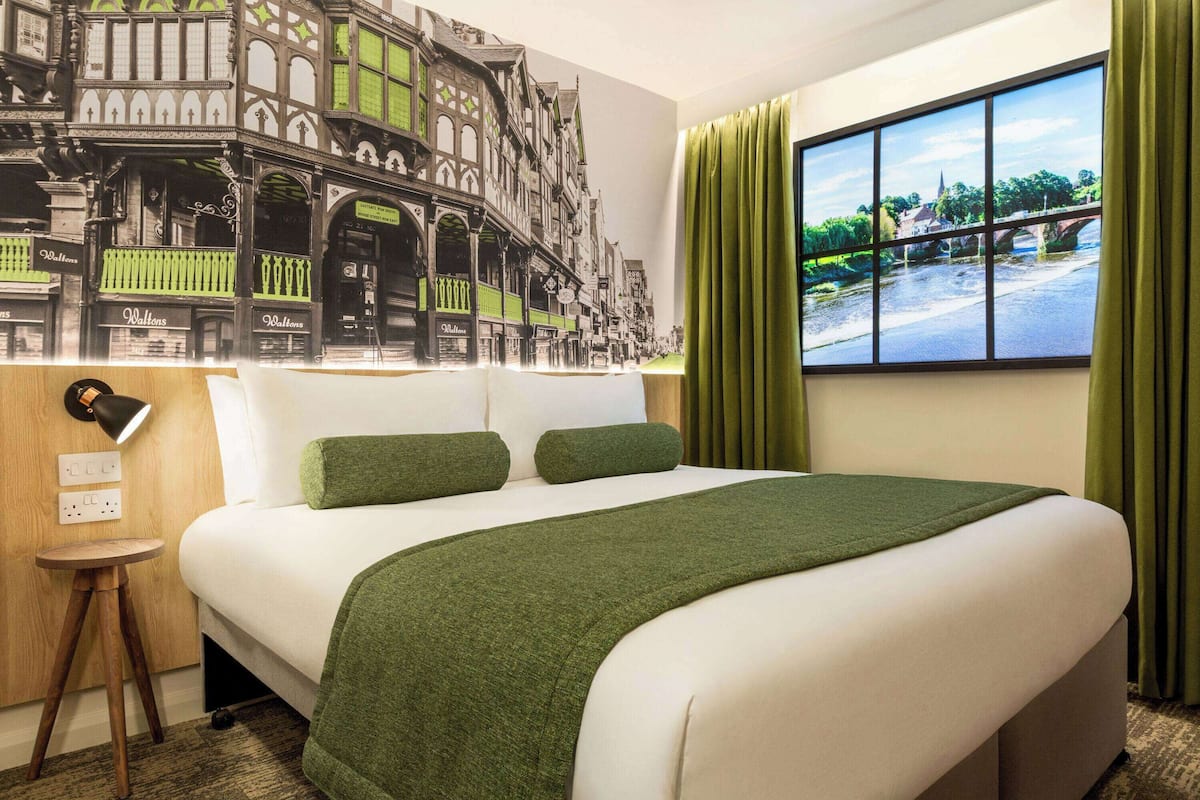 A hotel room with a large bed, green bedding, a mural of a street scene, a window with a view of a river and bridge, and a small wooden side table with a lamp.