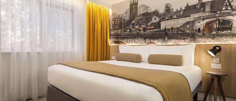 A hotel room with a large bed, a window with sheer curtains, a yellow curtain, a wall-mounted lamp, and a mural of a town scene.