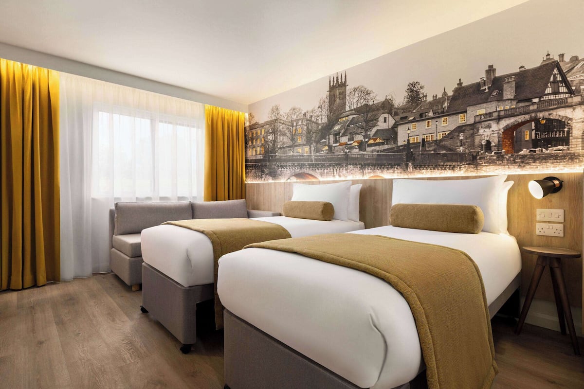 A hotel room with two beds, a sofa, a small table, and a mural of a town scene.