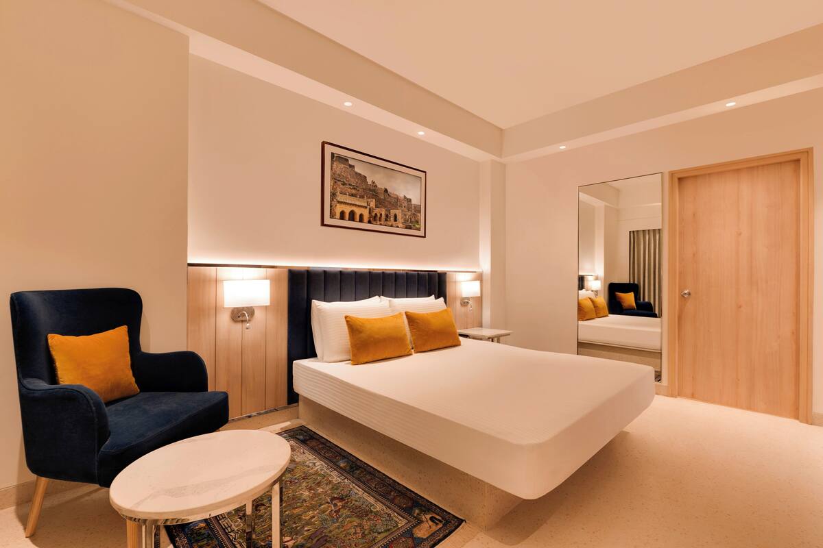 A modern hotel room with a large bed, a dark blue armchair, a small round table, and a large mirror.