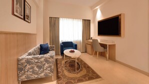 Ein kompaktes Hotelzimmer mit einer gemusterten Couch, einem kleinen runden Tisch, einem Schreibtisch, einem Stuhl und einem an der Wand befestigten Fernseher.