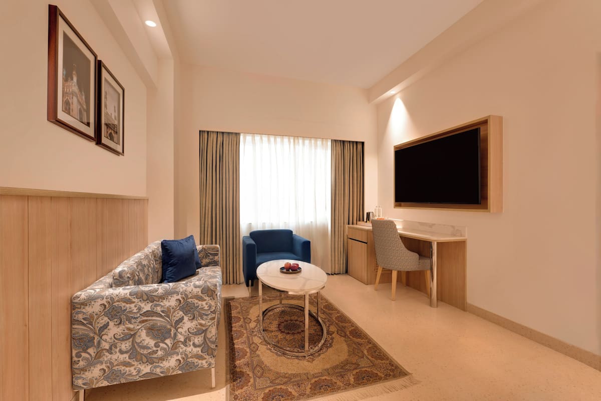 A compact hotel room with a patterned sofa, a small round table, a desk, a chair, and a television mounted on the wall.