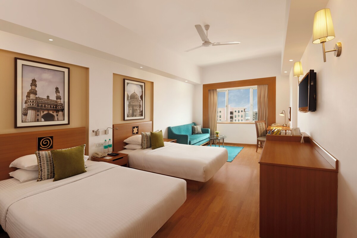 A hotel room with two beds, a desk, a chair, a small table, a TV, and framed pictures on the walls.
