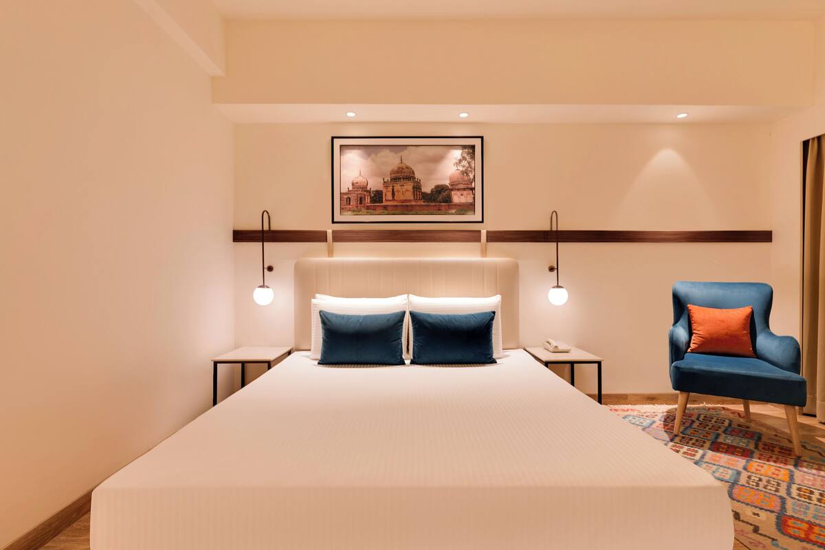 A hotel room with a large bed, two bedside tables, a blue armchair, a framed picture on the wall, and a patterned rug.