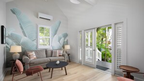 A modern living room with a sofa, two armchairs, a coffee table, and a small table with a lamp. There is a large window with shutters, an air conditioning unit, and a view of greenery outside. - Paradise Inn Key West - Adults Only (Key West)