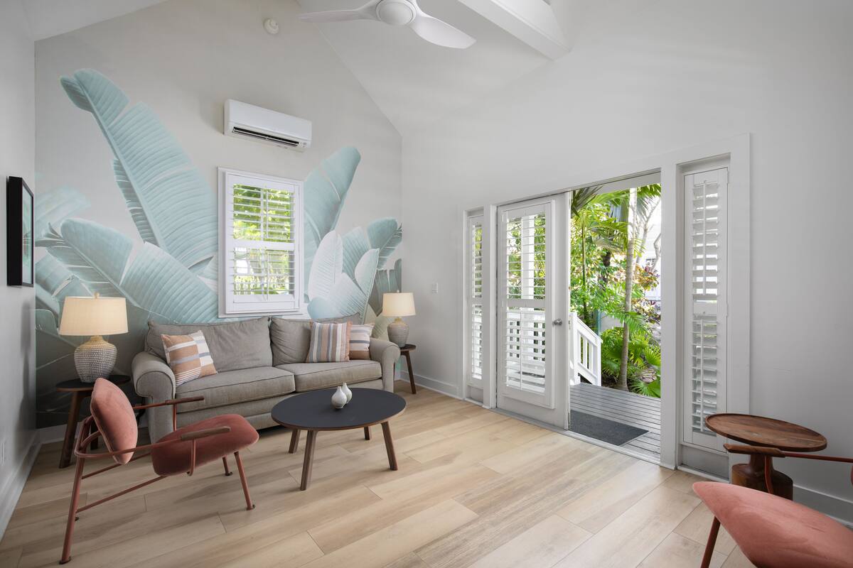 A modern living room with a sofa, two armchairs, a coffee table, and a small table with a lamp. There is a large window with shutters, an air conditioning unit, and a view of greenery outside.