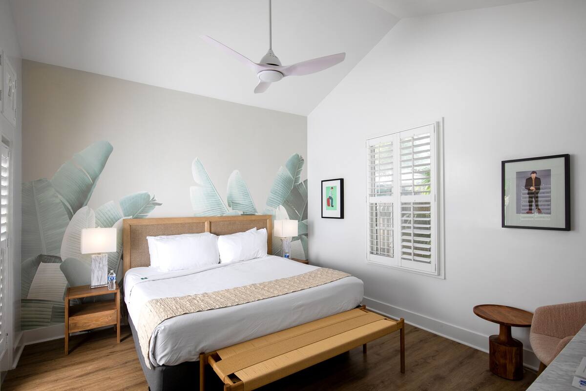 A bedroom with a bed, bedside tables, a ceiling fan, a bench, a chair, and a window with blinds.