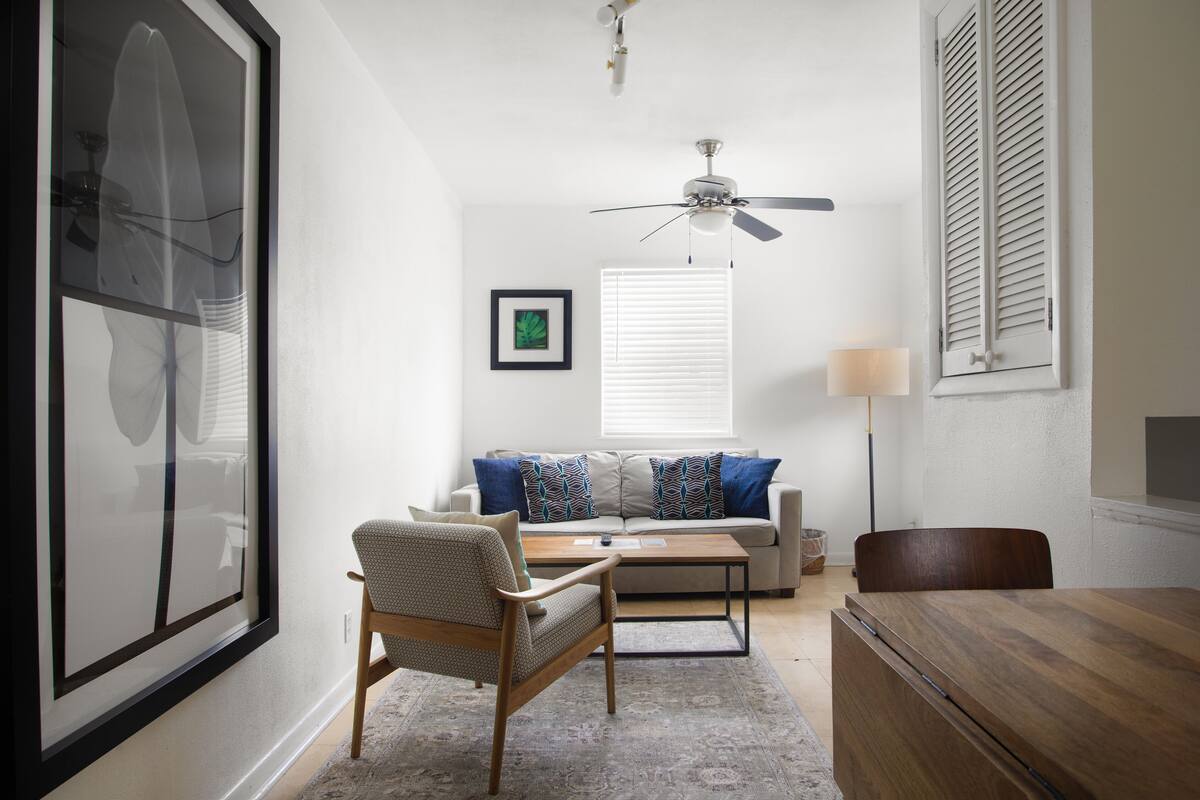 A living room with a sofa, two chairs, a coffee table, a ceiling fan, and a window with blinds.