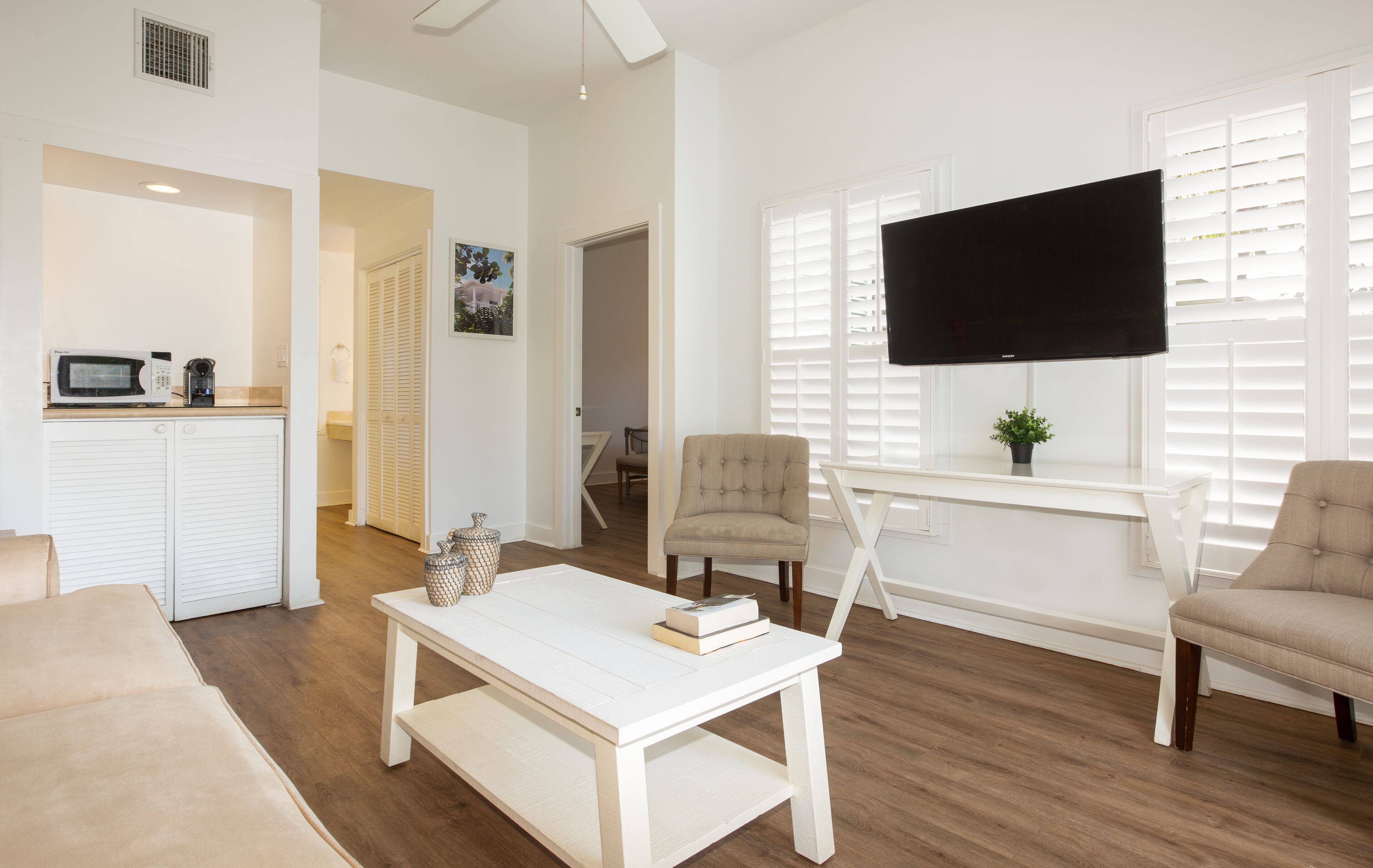 Un salon moderne avec un canapé blanc, une table basse sur laquelle repose un livre, un fauteuil beige, un téléviseur posé sur un meuble blanc, et un coin cuisine équipé d’un four à micro-ondes et d’une machine à café.