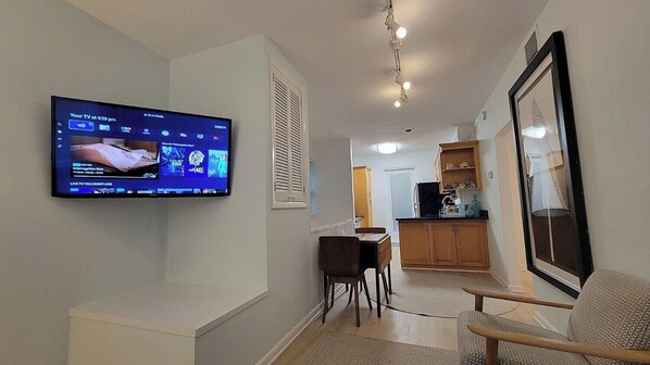 A living room with a flat-screen TV mounted on the wall, a dining area with a table and chairs, and a kitchen area with cabinets and a countertop.