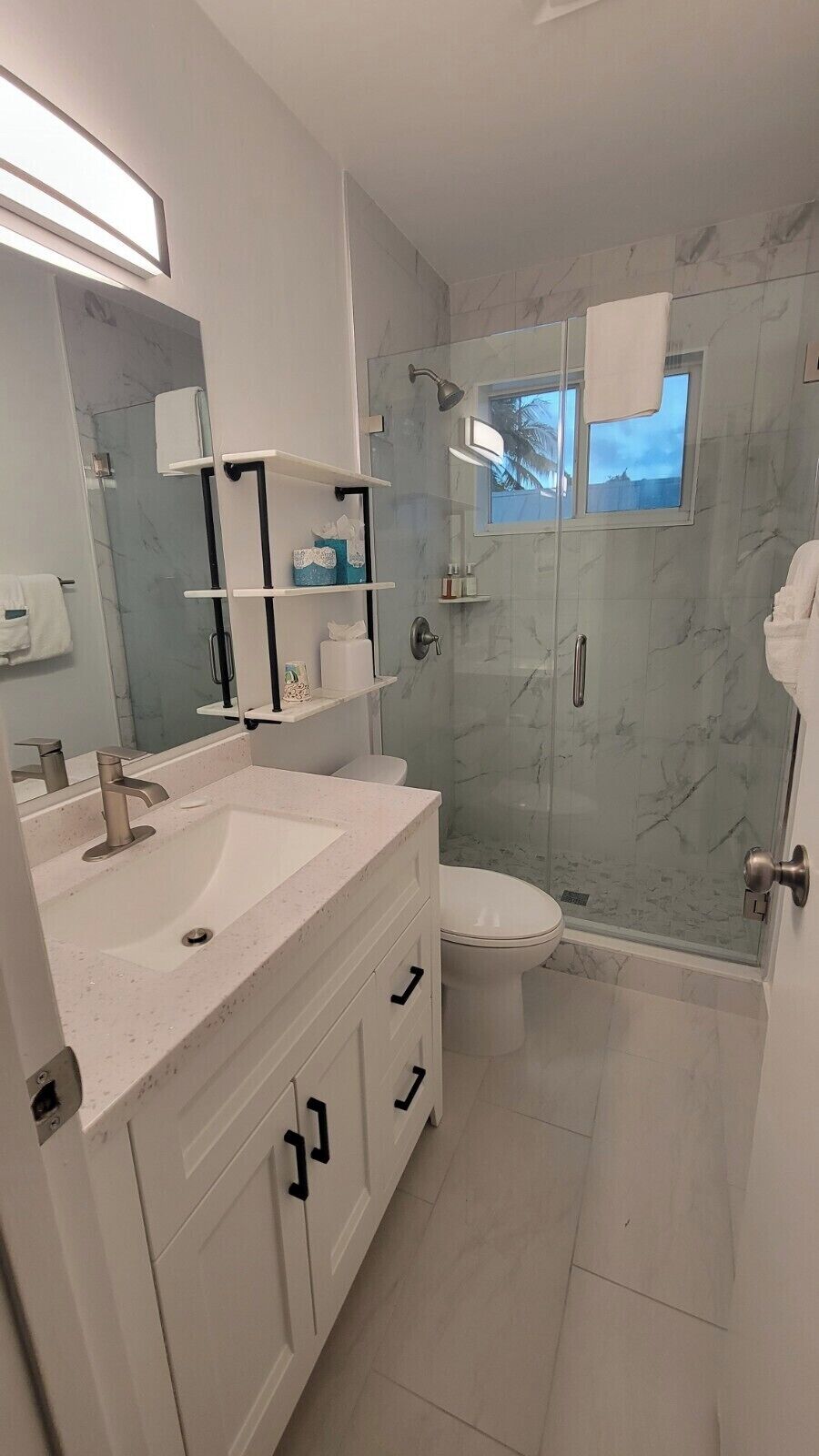 Un baño moderno con ducha de mármol, inodoro, lavamanos con espejo y estantes con artículos de aseo.