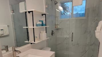 Un baño moderno con ducha de mármol, inodoro, lavamanos con espejo y estantes con artículos de aseo.