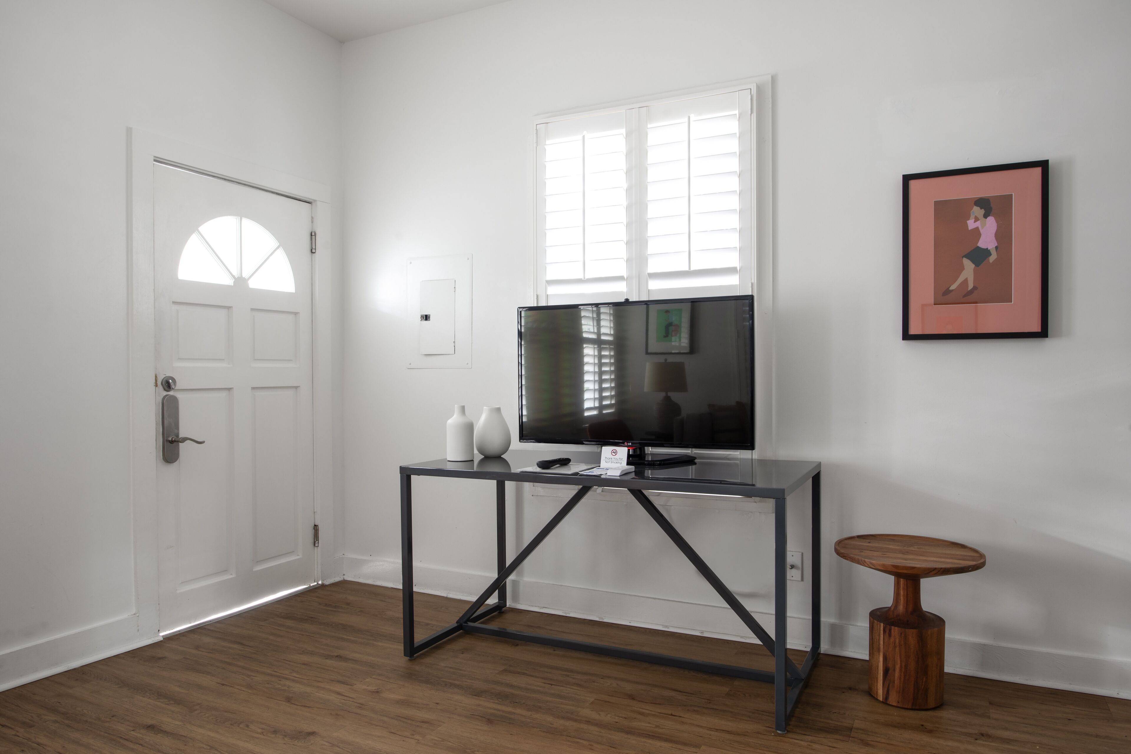A room with a wooden floor, a white door, a glass TV stand with a flat-screen TV, two white vases, and a framed picture on the wall.