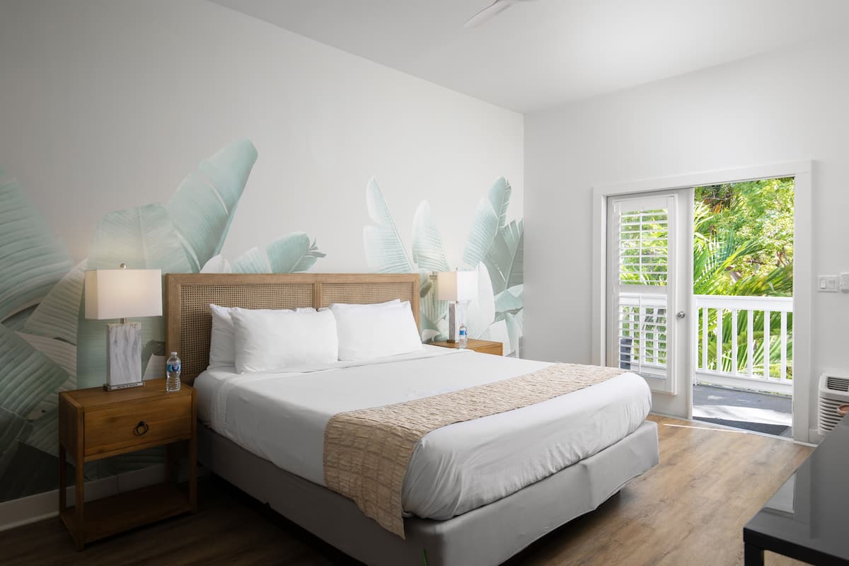 A bedroom with a large bed, wooden headboard, two bedside tables with lamps, a view of greenery through a sliding door, and a balcony.