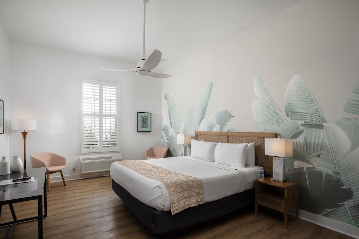 A hotel room with a bed, a desk, a chair, a standing lamp, a ceiling fan, and a large window with blinds.