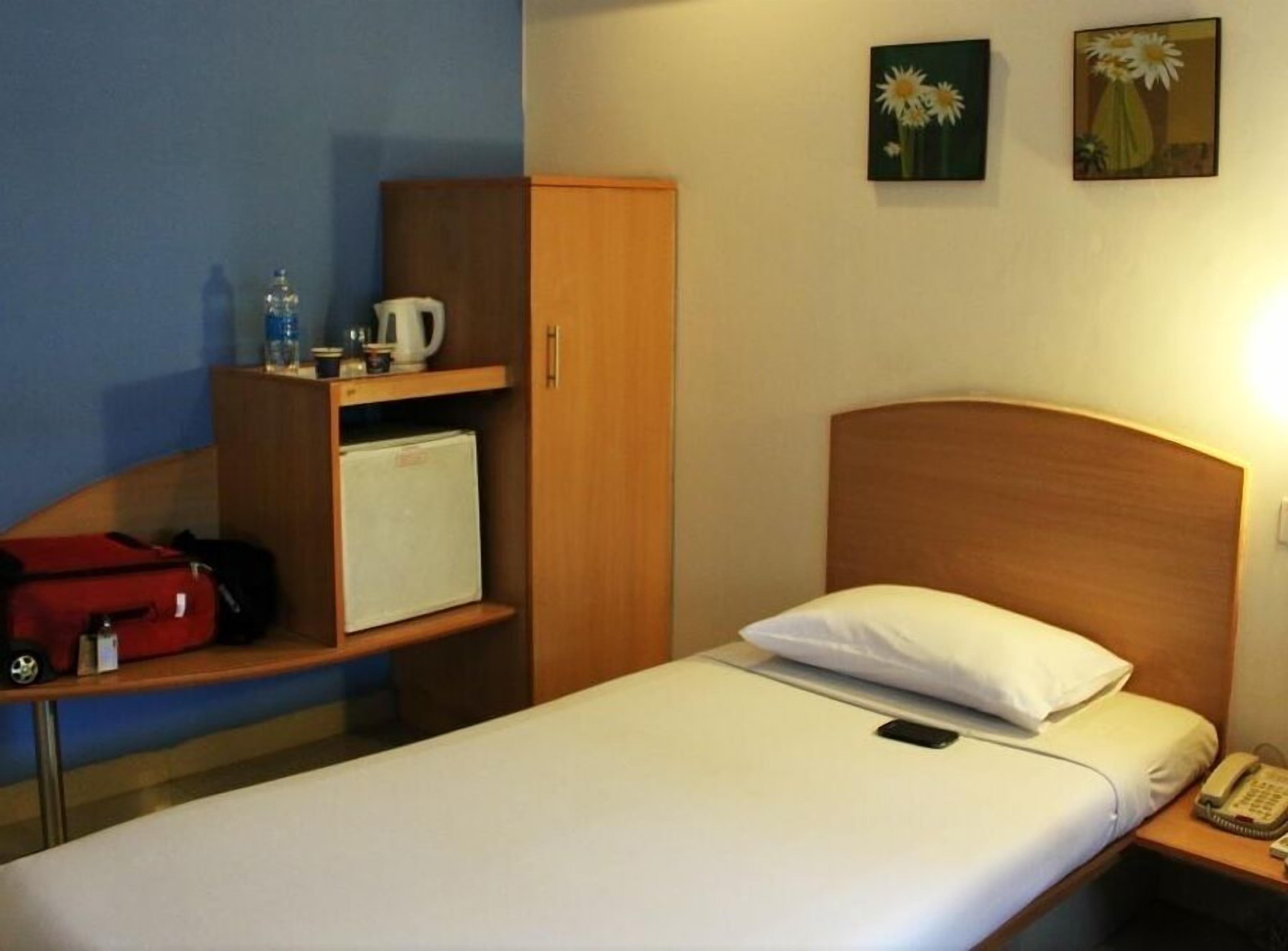 A hotel room with a single bed, a wooden headboard, a nightstand with a phone, a small cabinet with a kettle and a red bag, and two flower paintings on the wall.