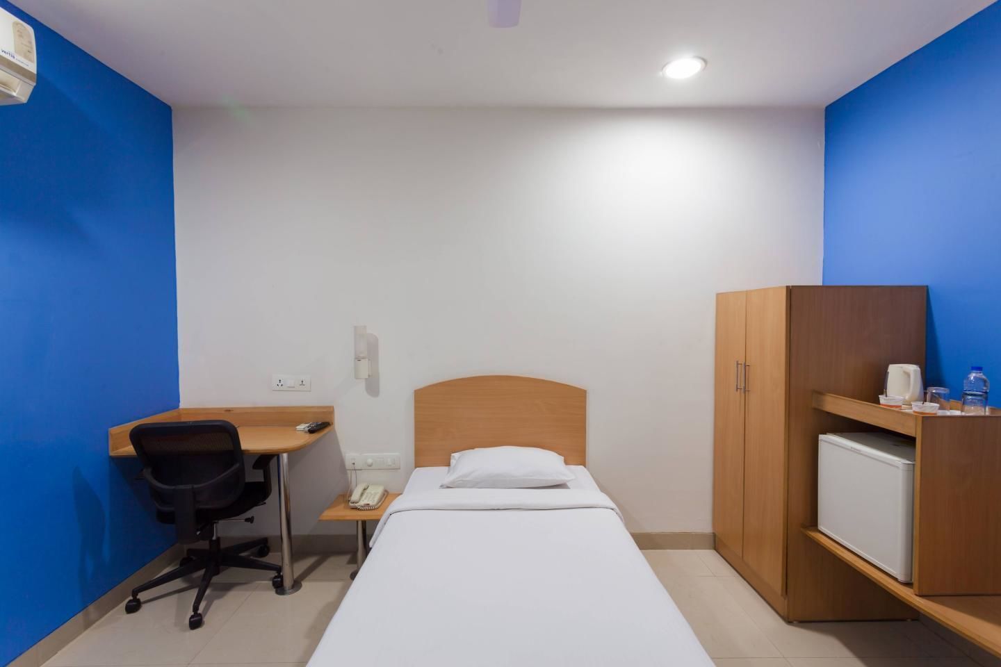 A small, single-bed room with a wooden desk, a chair, a wardrobe, and a mini-fridge.