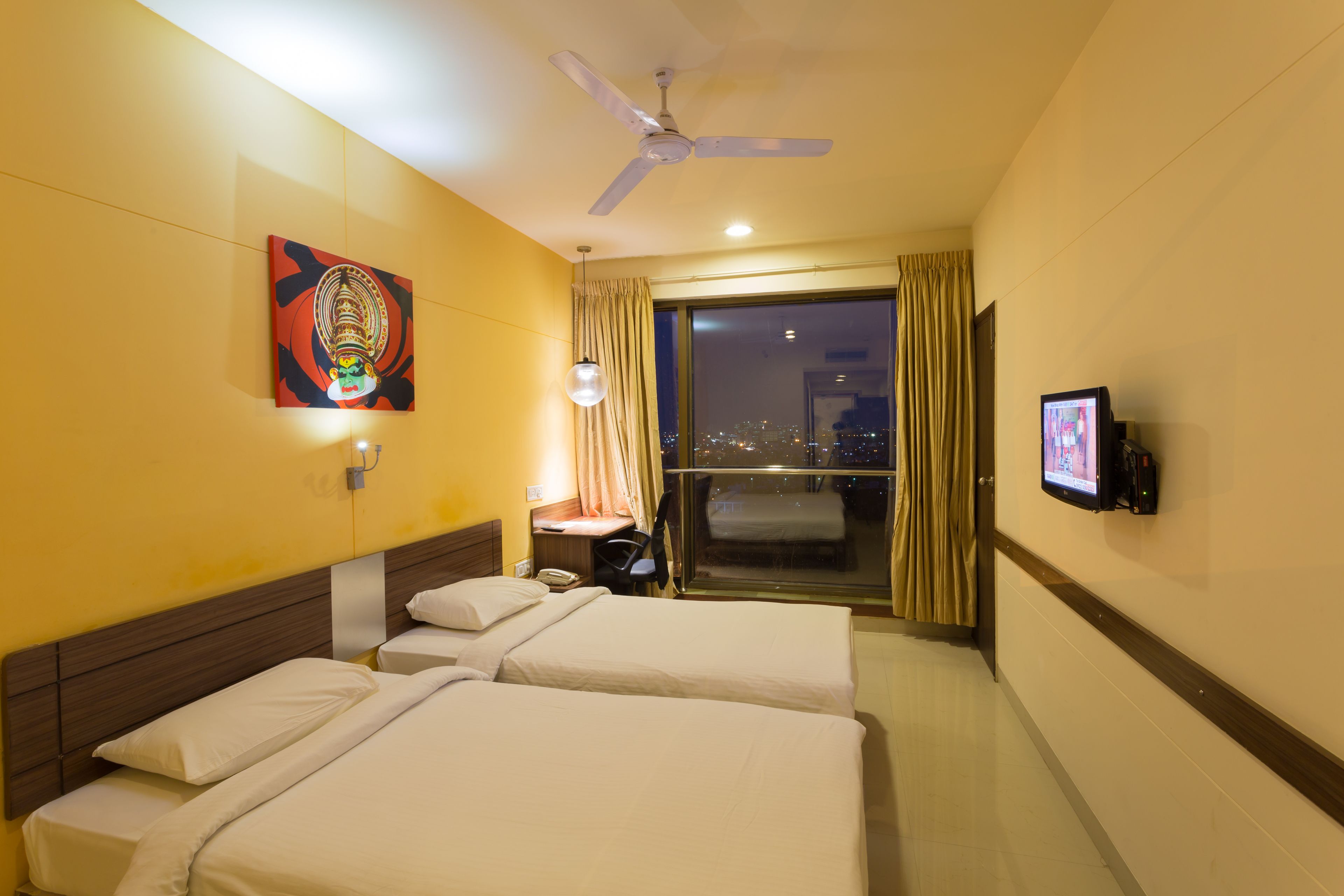 Habitación de hotel con dos camas, ventilador de techo, mesita de noche con lámpara, televisión y un ventanal grande con cortinas.