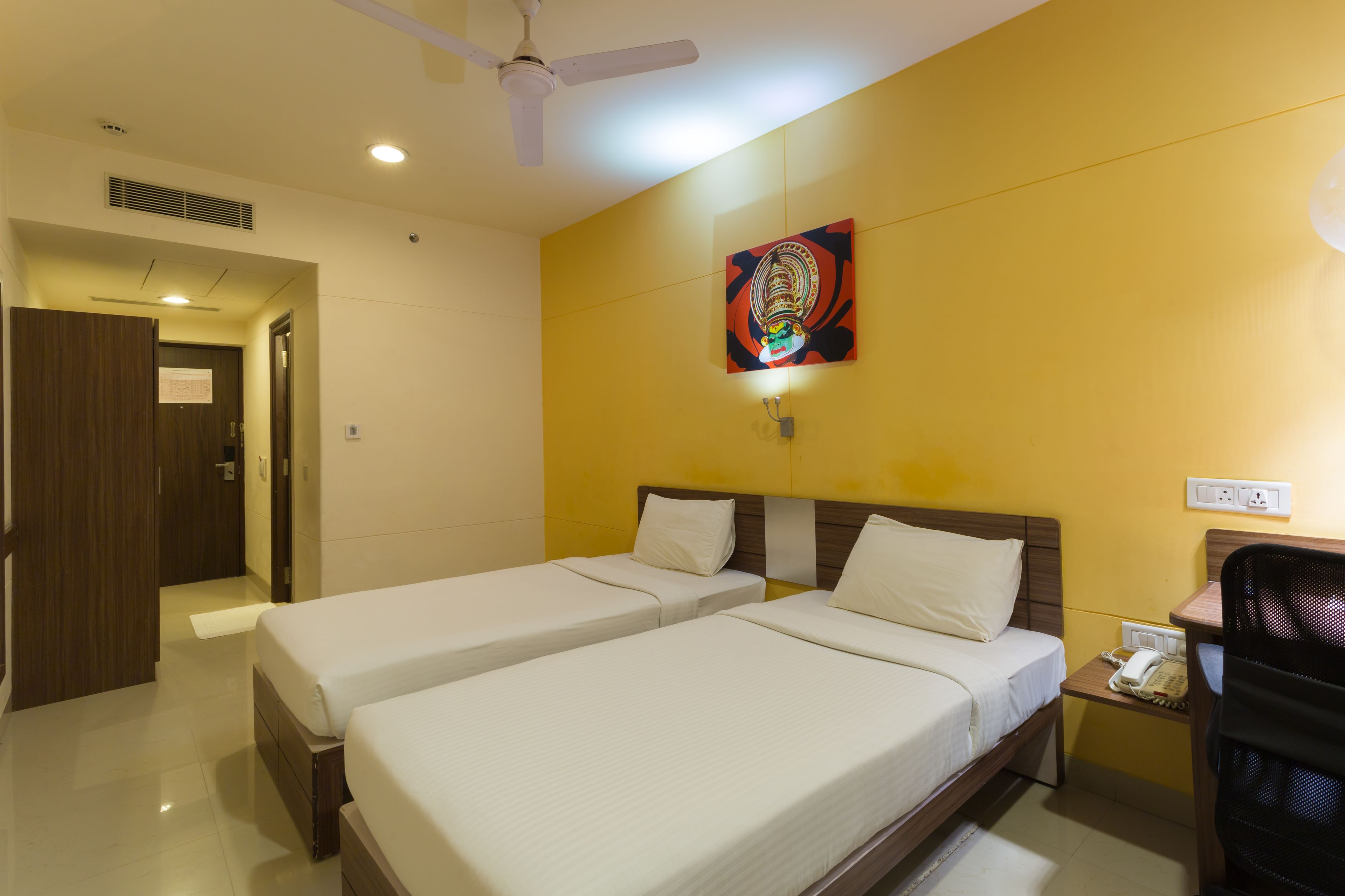 A hotel room with two single beds, a desk with a telephone, a chair, a ceiling fan, and a painting on the wall.