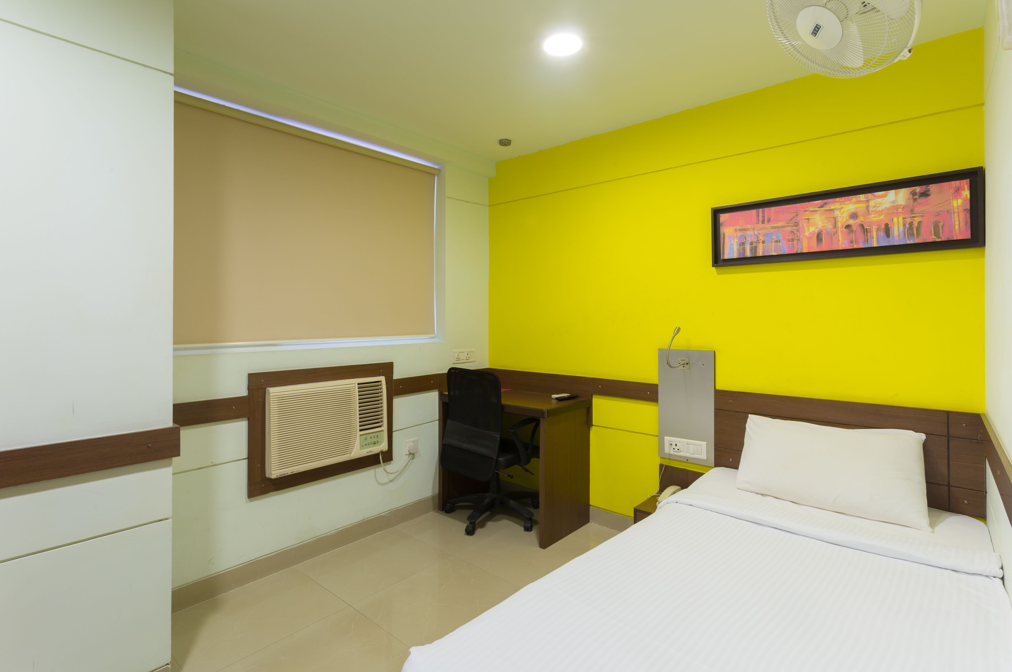 A hotel room with a bed, a desk, a chair, a window with blinds, a wall-mounted air conditioner, and a ceiling fan.