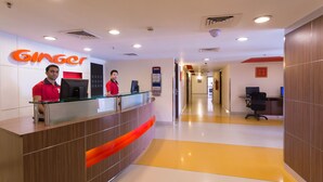 A reception area with a modern design, featuring a reception desk with a glass top and a wooden finish. There are two receptionists in red uniforms, and a digital screen is visible on the desk. The floor has a yellow and white color scheme, and the walls are adorned with a large orange sign reading "Ginger." - Ginger Chennai (Chennai)