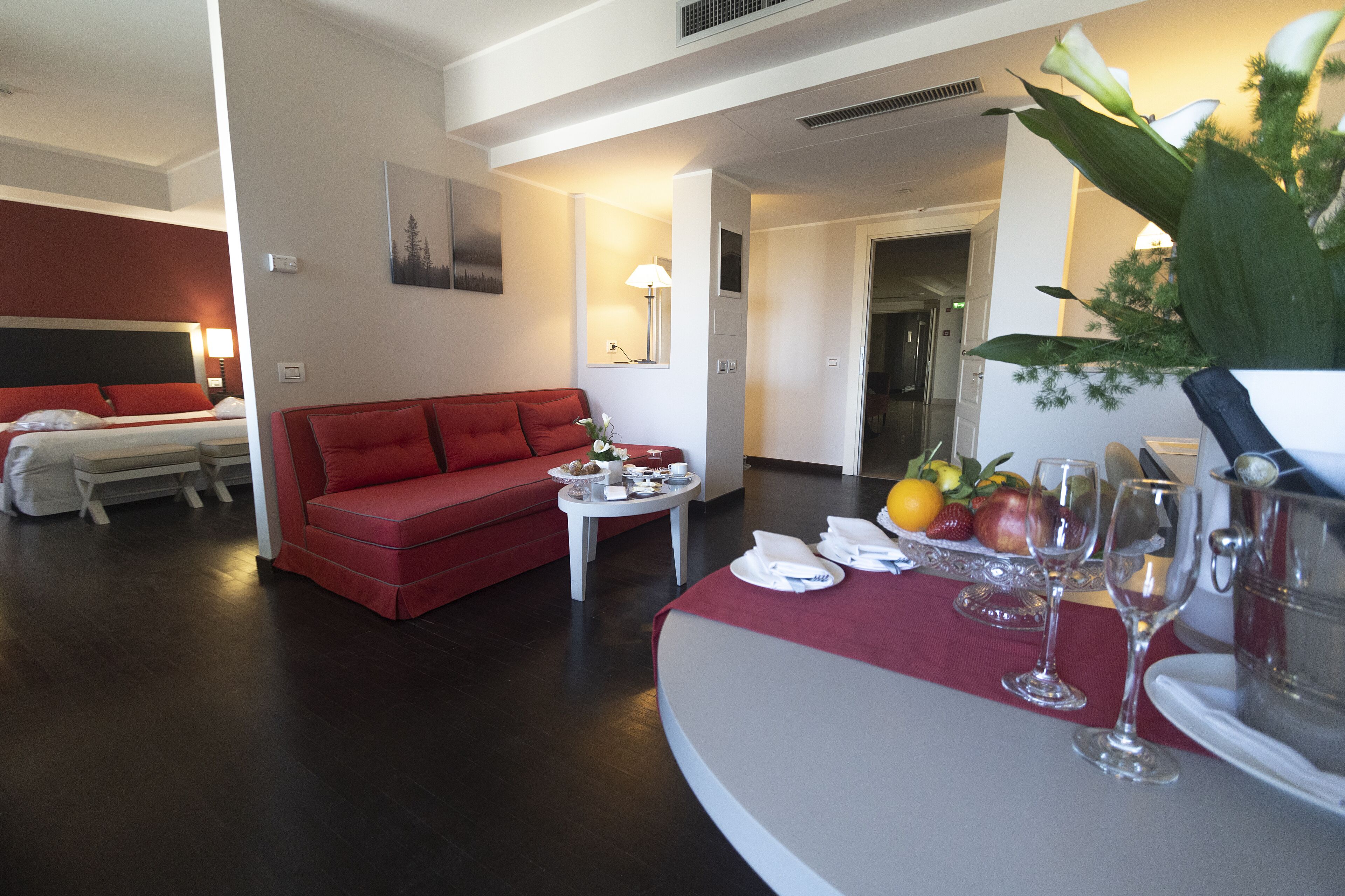 Habitación de hotel con cama, sofá rojo, mesa de comedor con fruta y copas de vino, y una planta grande.