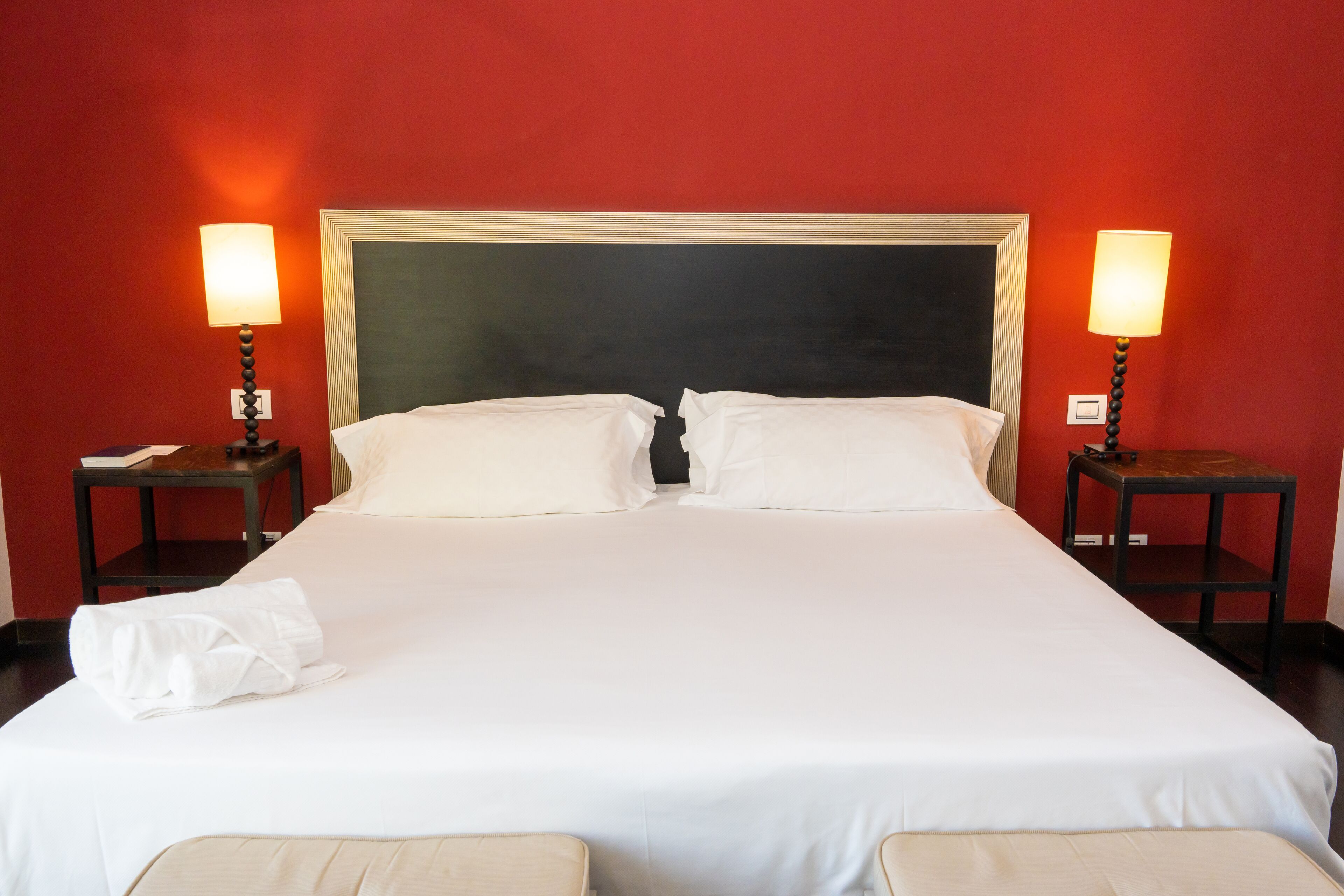 A neatly made double bed with white linens, two bedside tables with lamps, and a red wall.