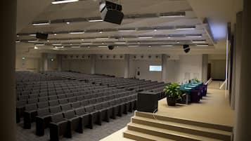 Um grande auditório com assentos em níveis, palco com um pódio e uma longa mesa com cadeiras.