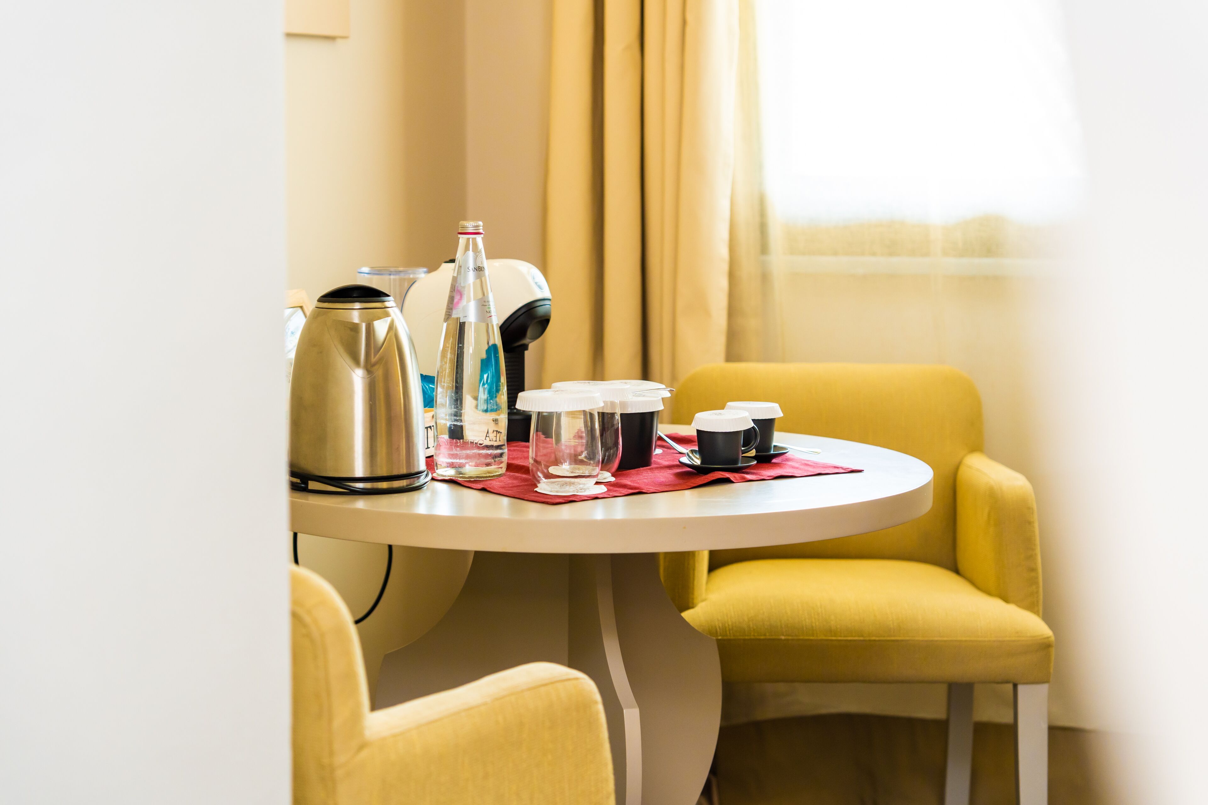 A hotel room with a round dining table, yellow chairs, a coffee maker, a bottle, and a glass.