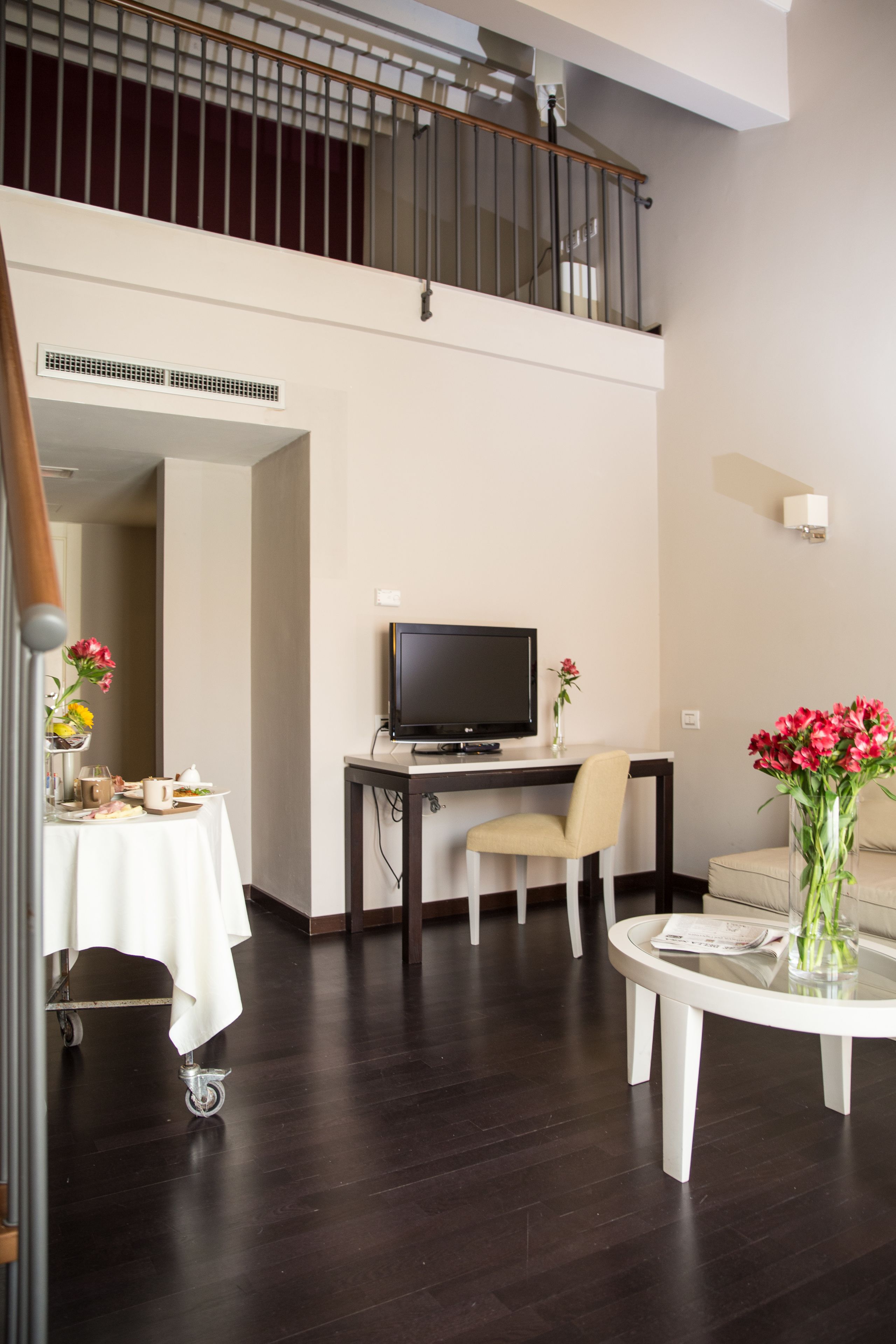 A modern hotel room with a dining area, a flat-screen TV, a round table, and a balcony with a railing.