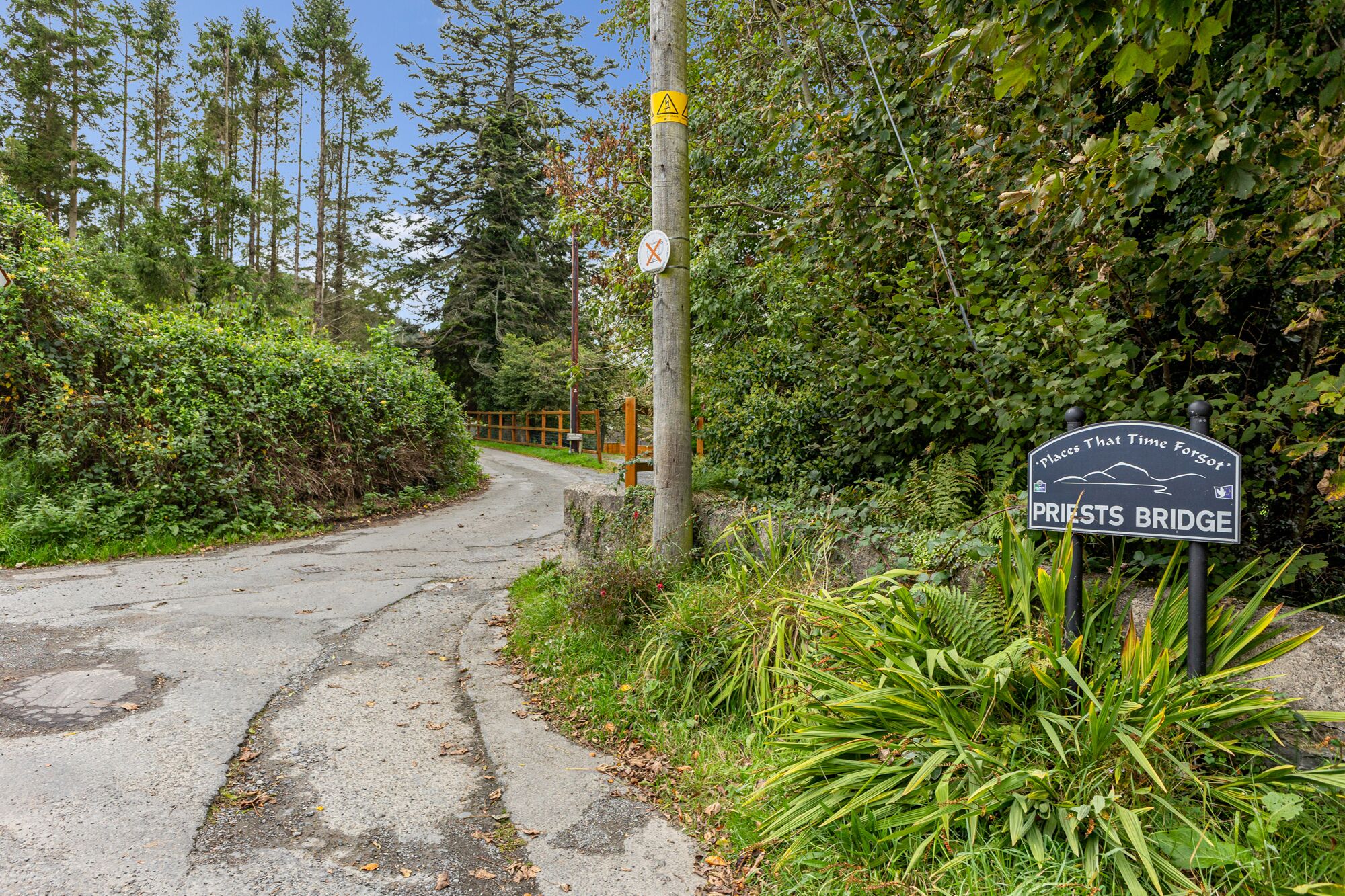 A rural road leading to a bridge with a sign indicating 'Places That Time Forgot' and 'Priests Bridge'.