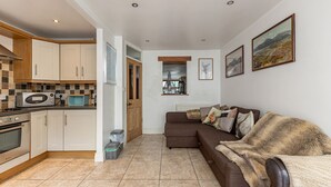 A modern kitchen with white cabinets, a microwave, and a stove. There is a brown sofa with a fur throw and pillows. A window with a view of mountains is visible. - Priest Bridge Cottage (Newcastle)