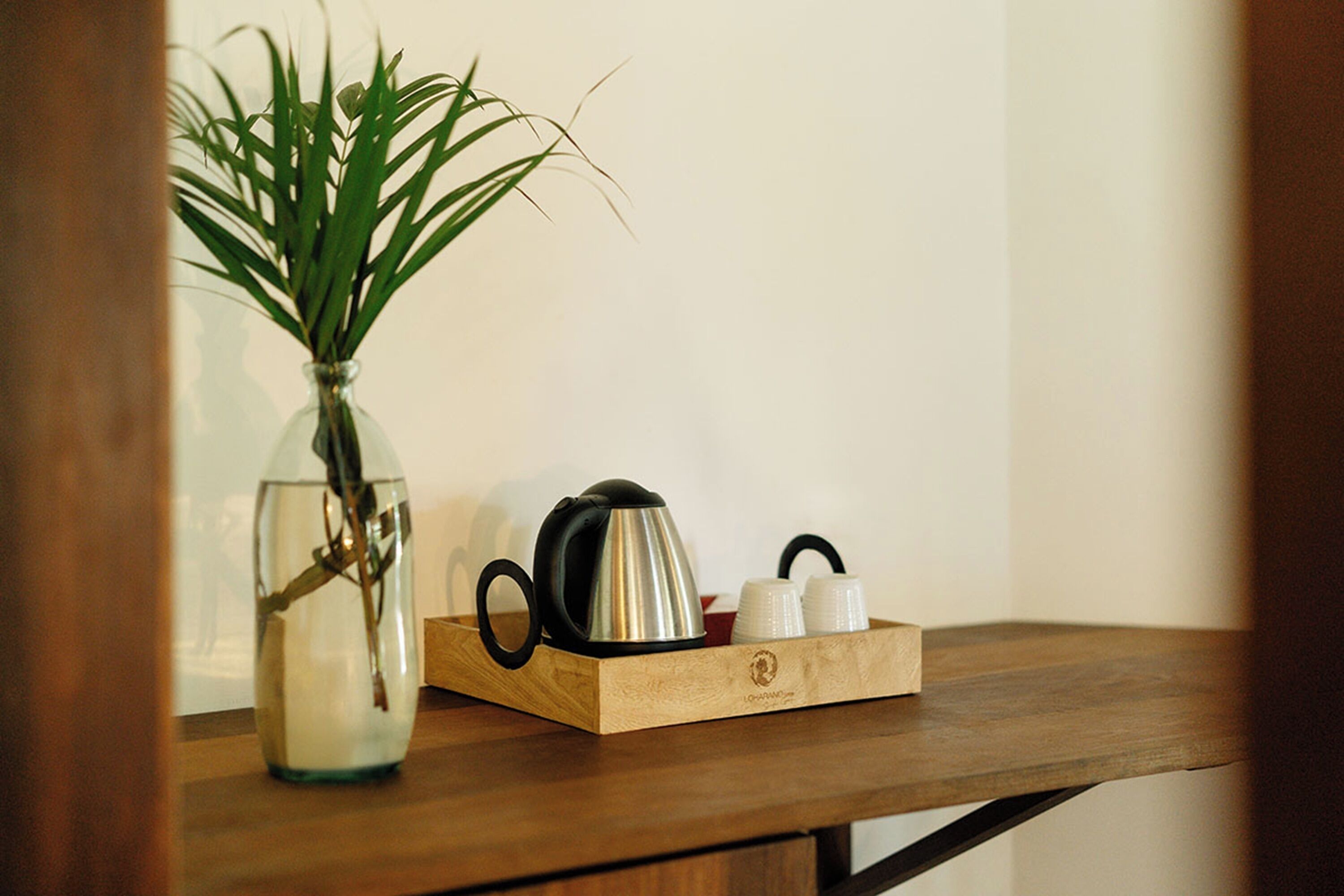 Une table en bois sur laquelle se trouvent un vase en verre contenant une plante, une bouilloire noire et deux tasses blanches disposées sur un plateau.